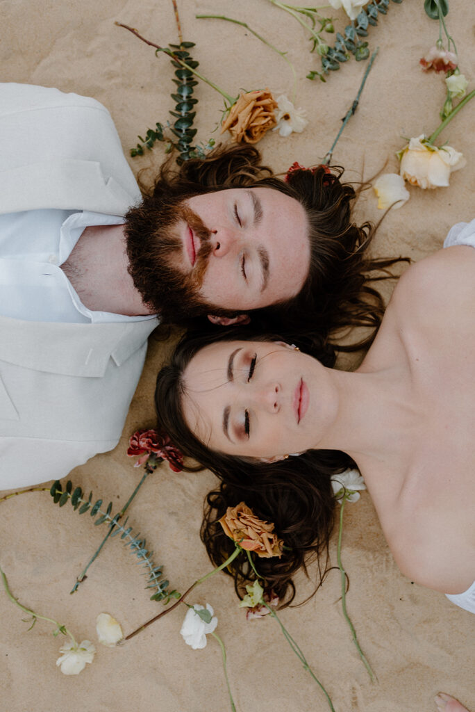 couple laying down in the sand surrounded by loose flowers